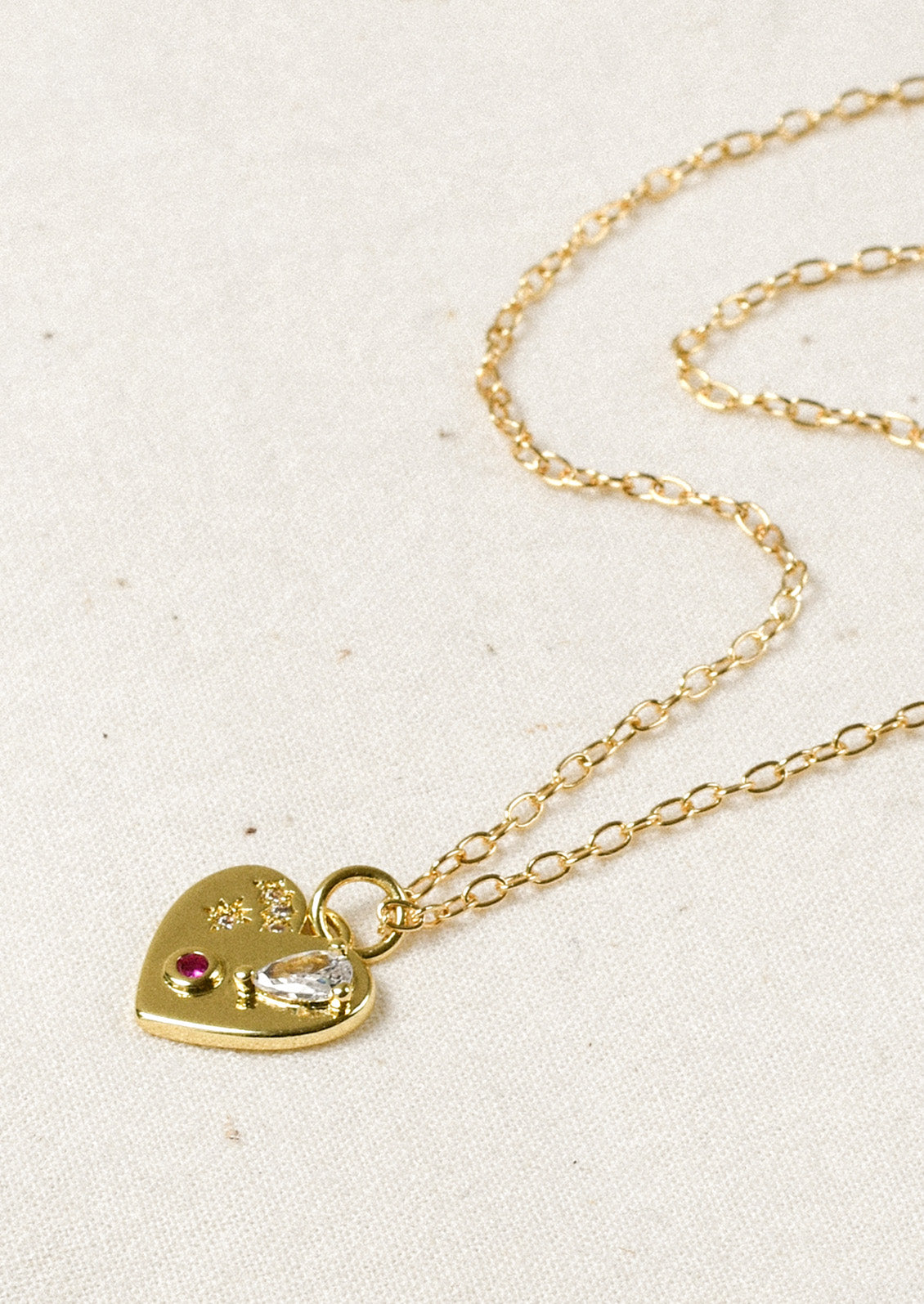A gold necklace with a gold heart charm adorned with glass crystal and a red gemstone.