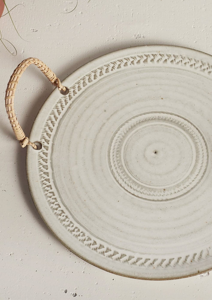 A round ceramic tray with neutral finish and rattan handles.