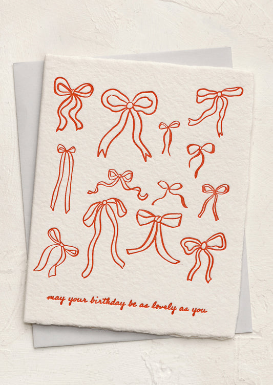A greeting card with allover red bow print, text reads "May your birthday be as lovely as you".