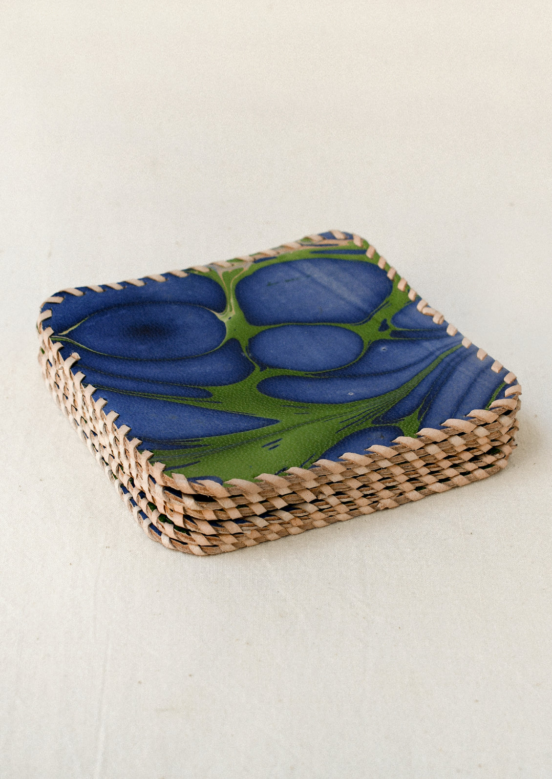 A stack of square leather coasters with rounded corners, featuring an abstract marbled pattern in deep indigo blue and green. The edges are finished with contrasting cream-colored whipstitching.