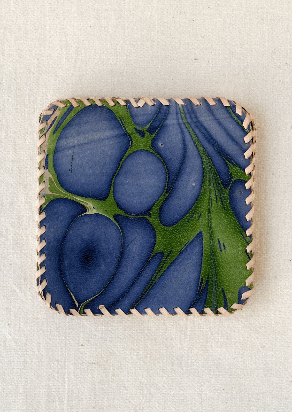 Square leather coaster with rounded corners, featuring an abstract marbled pattern in deep indigo blue and green. The edges are finished with contrasting cream-colored whipstitching.