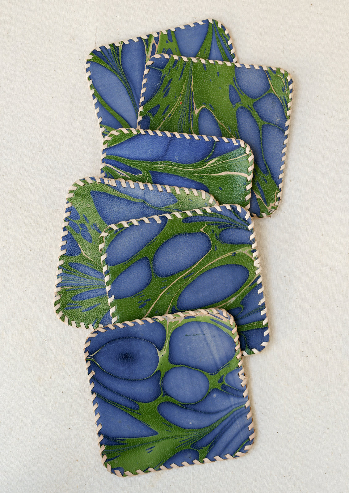 Square leather coasters with rounded corners, featuring an abstract marbled pattern in deep indigo blue and green. The edges are finished with contrasting cream-colored whipstitching.