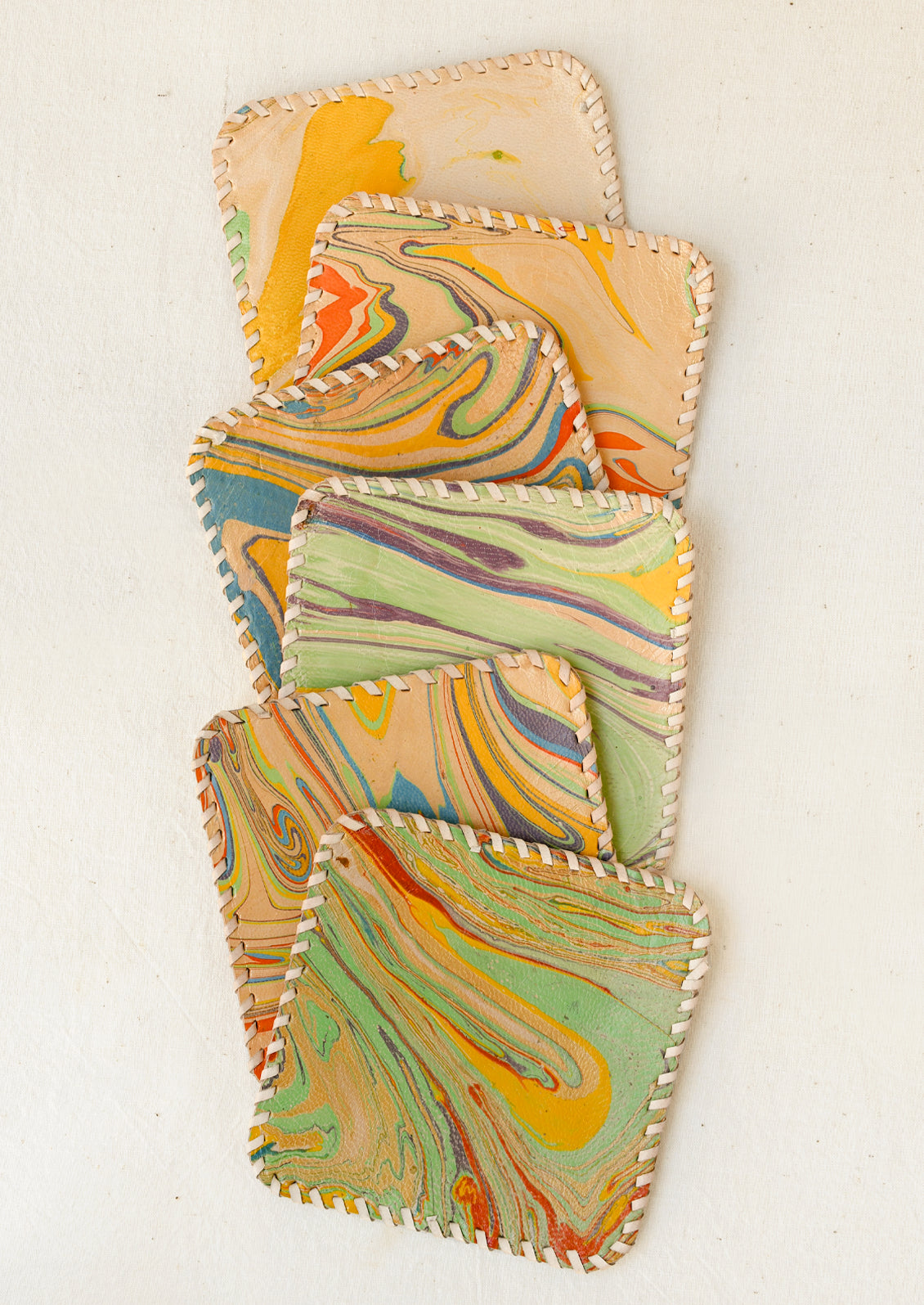 Square leather coasters with rounded corners, featuring a colorful swirling marbled pattern in mustard yellow, green, blue, orange, and purple. The edges are finished with contrasting cream-colored whipstitching.