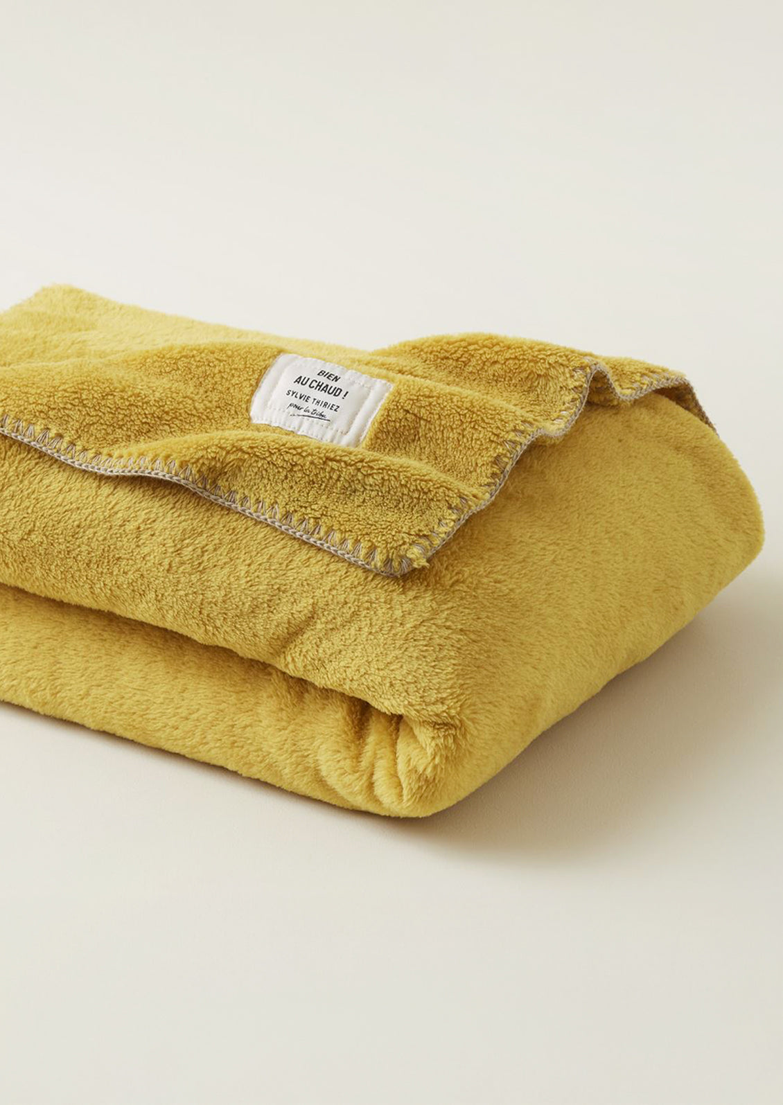 A folded mustard yellow fleece blanket with a white woven label and a light beige stitched border.