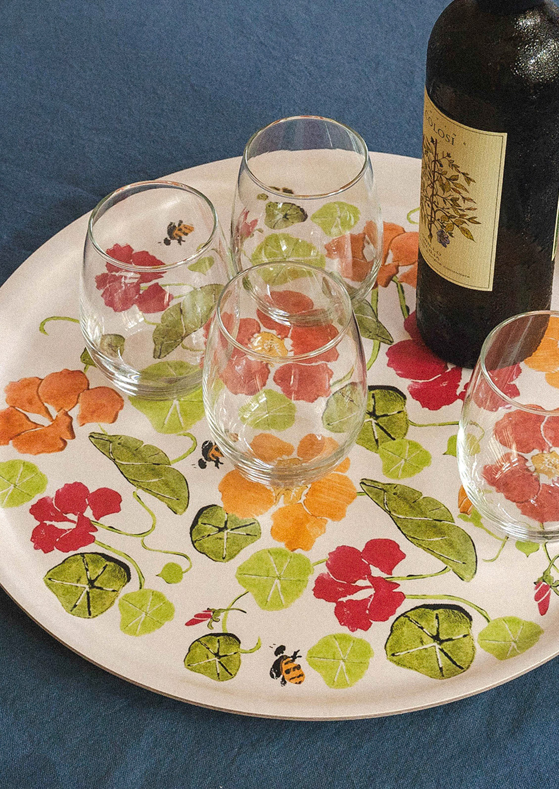 A wooden circle tray with an illustration of nasturtium flowers and bees on a table with a blue tablecloth. Wine glasses and a wine bottle sit on top of the tray.