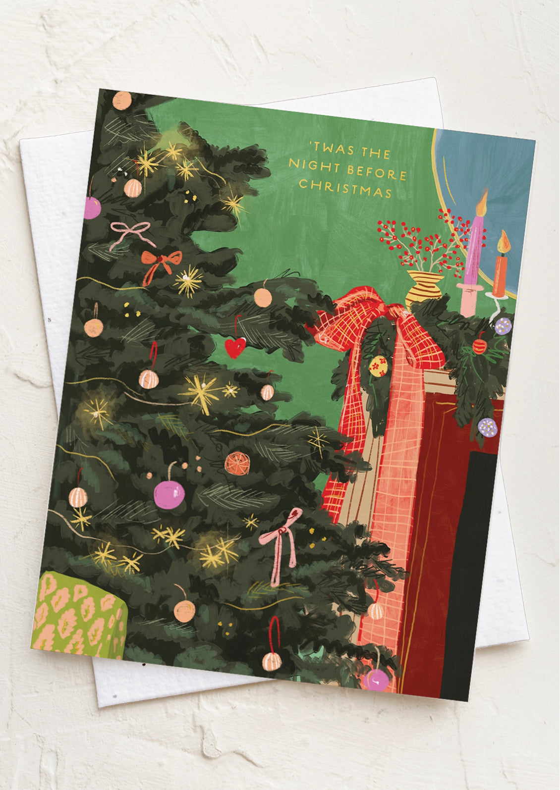 A Christmas greeting card featuring an illustrated green Christmas tree with ornaments, a red bow, candles, and wrapped gifts, layered over a white envelope on a light textured background. The card reads, 'Twas the Night Before Christmas.