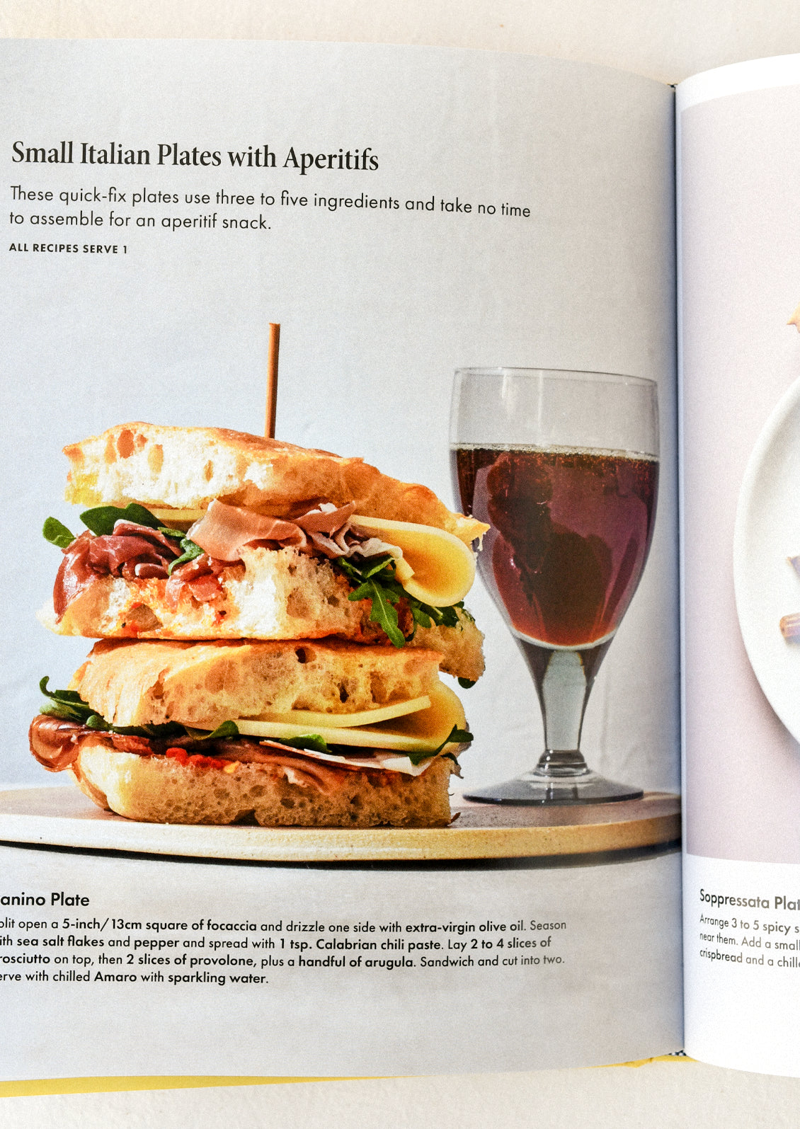 A cookbook open on a table with an image of a panini and a sparkling drink.