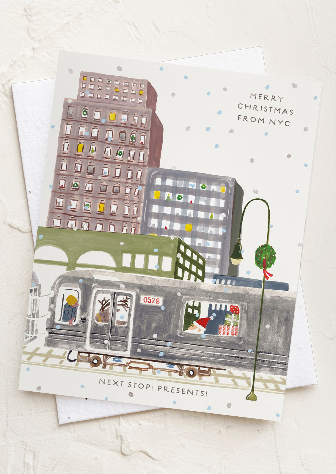A Christmas-themed greeting card featuring an illustrated city street scene with a subway train, buildings, and a lit lamp post, shown on a textured white background with a white envelope behind it.