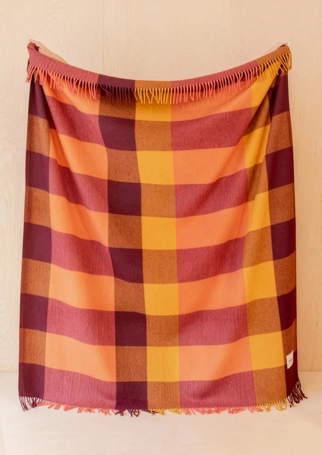 A plaid fringed throw blanket in warm colors, featuring stripes of orange, yellow, burgundy, and brown, hanging against a light wood-paneled wall.