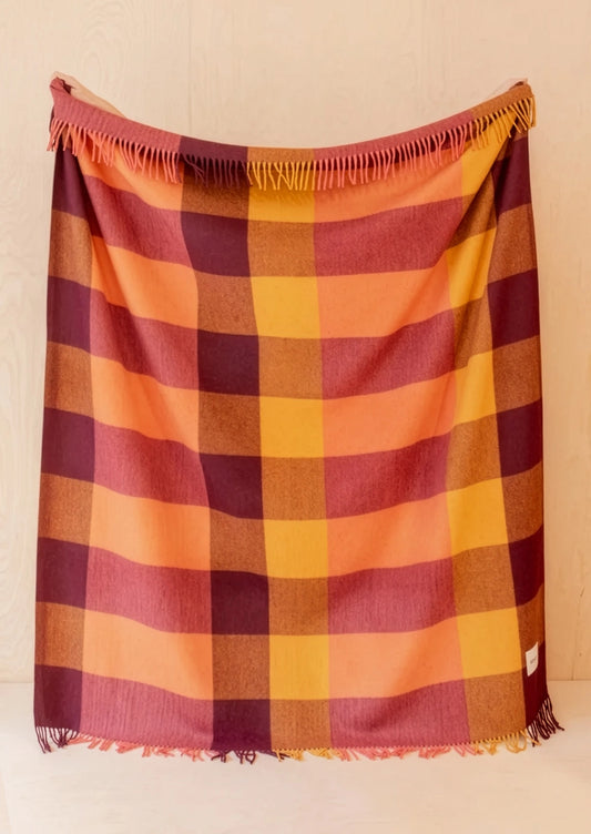 A plaid fringed throw blanket in warm colors, featuring stripes of orange, yellow, burgundy, and brown, hanging against a light wood-paneled wall.