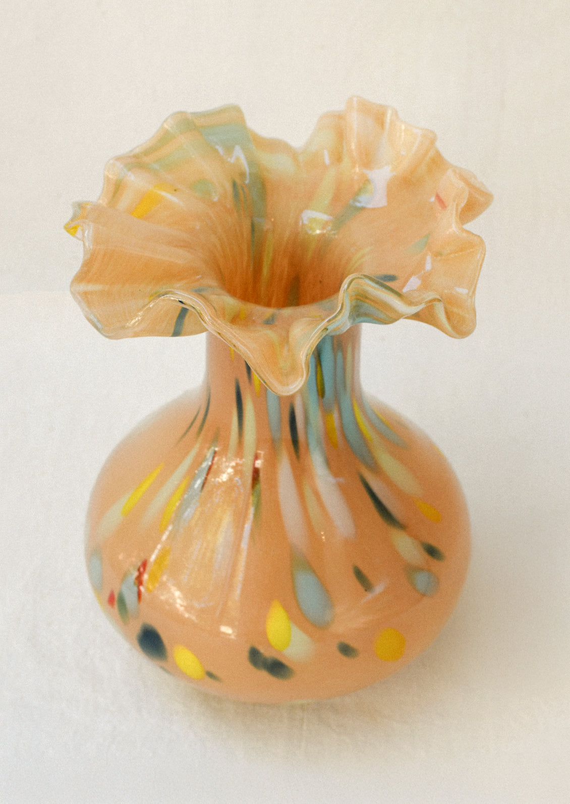 A handblown glass vase with a peach colored body, multicolor paint strokes, and a ruffled opening.