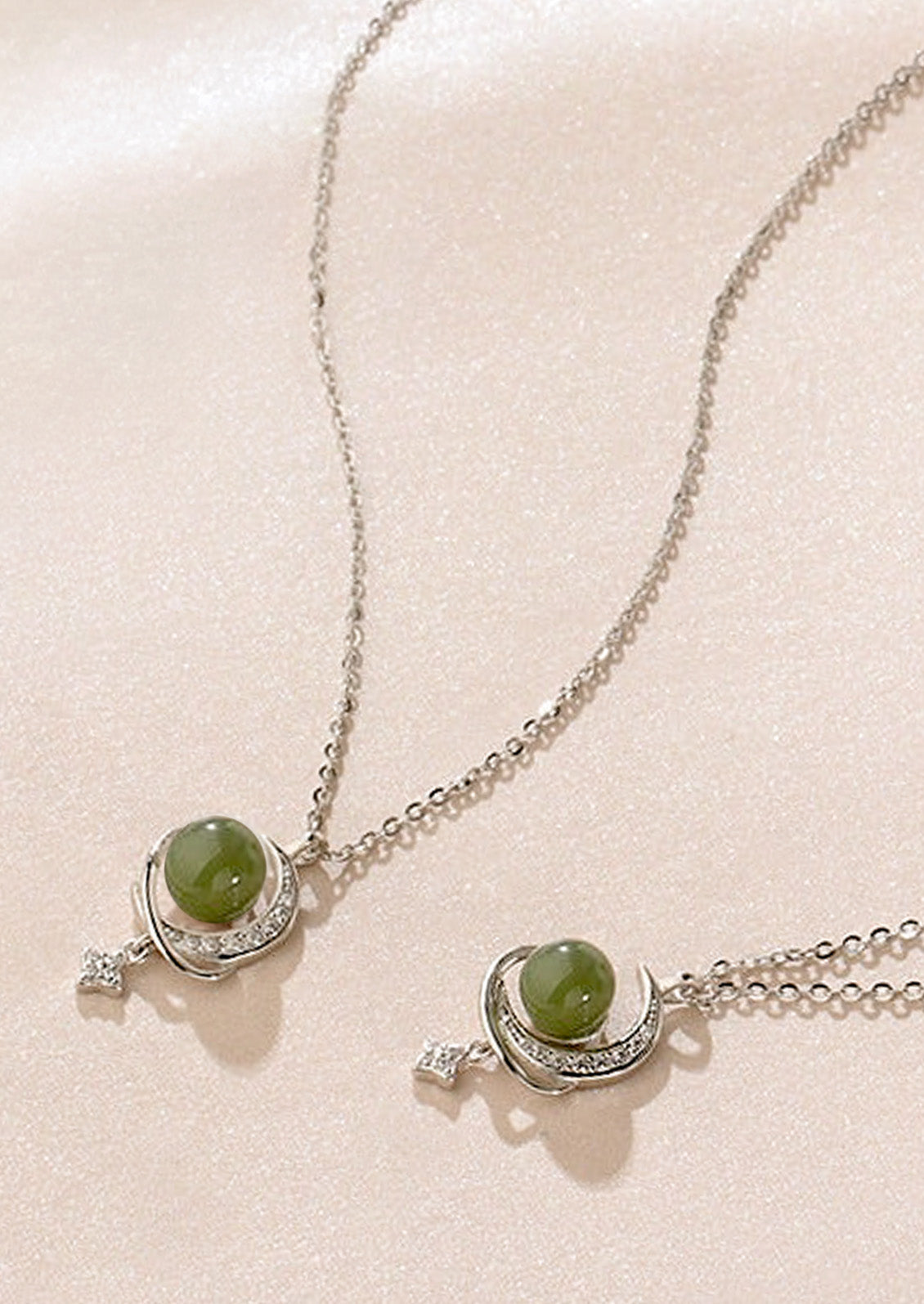 Two sterling silver necklaces with a moon and star charm holding a jade stone in the center laying on a pink background.