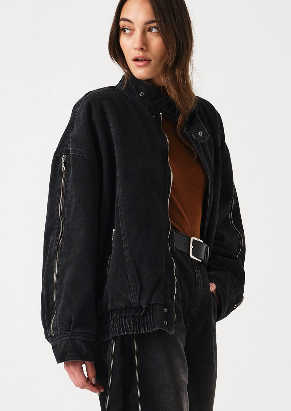 A woman wearing a black washed denim jacket with silver zippers, styled over a brown top and black pants, photographed against a light gray studio background.