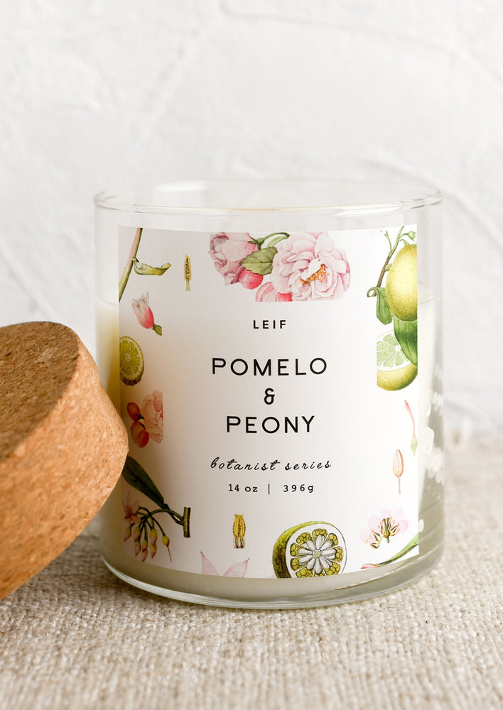 A glass candle with a cork lid and white botanical printed label reading "pomelo and peony".