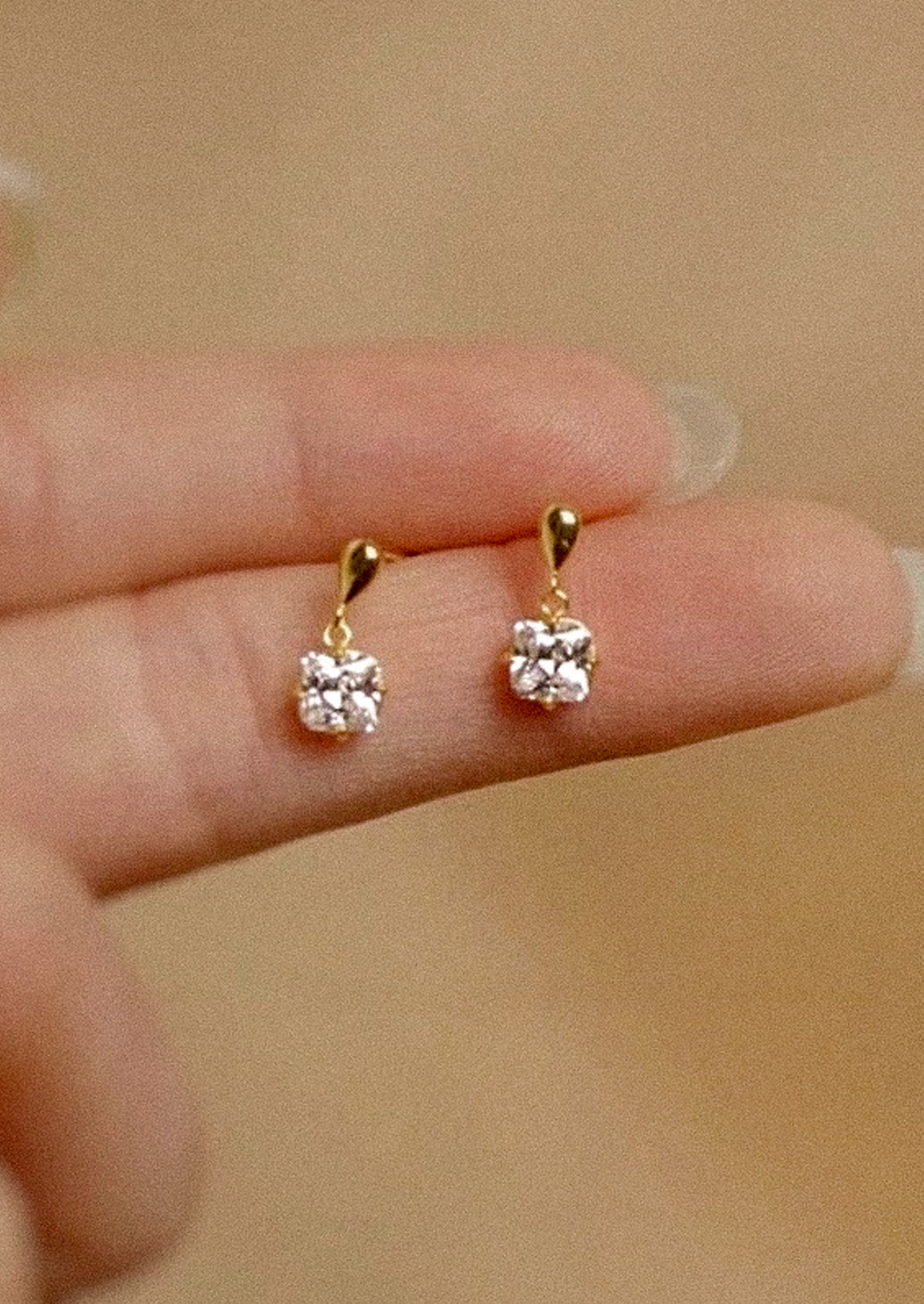 A woman holding pair of gold earrings with a princess cut cubic zirconia.