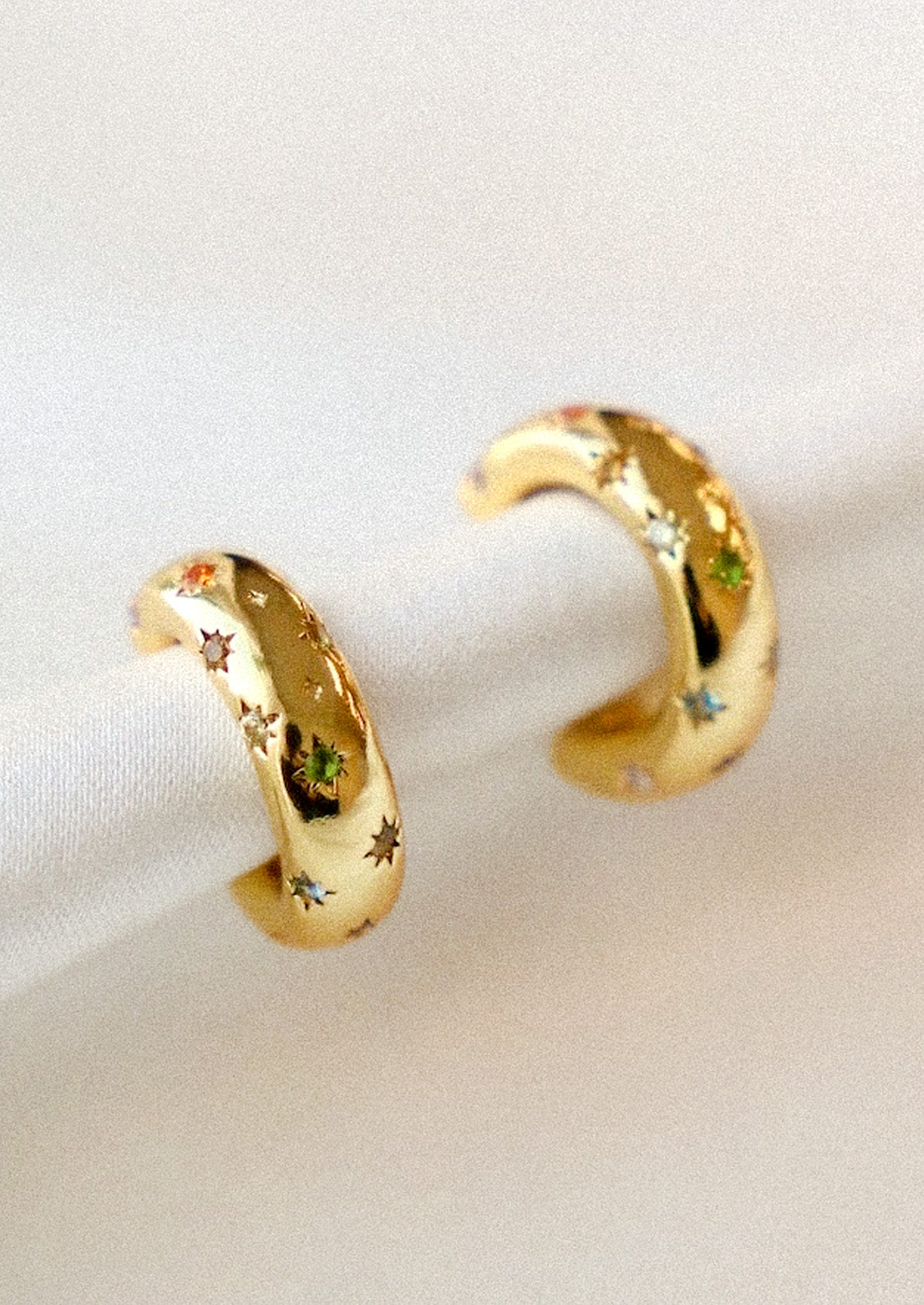 A pair of gold hoop earrings with small starbursts throughout adorned with various colored gems in the centers attached to a white fabric.