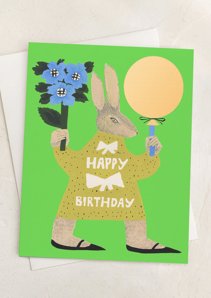 A green card with illustration of human-like rabbit holding flowers and balloon, text on rabbit's shirt reads "Happy Birthday".