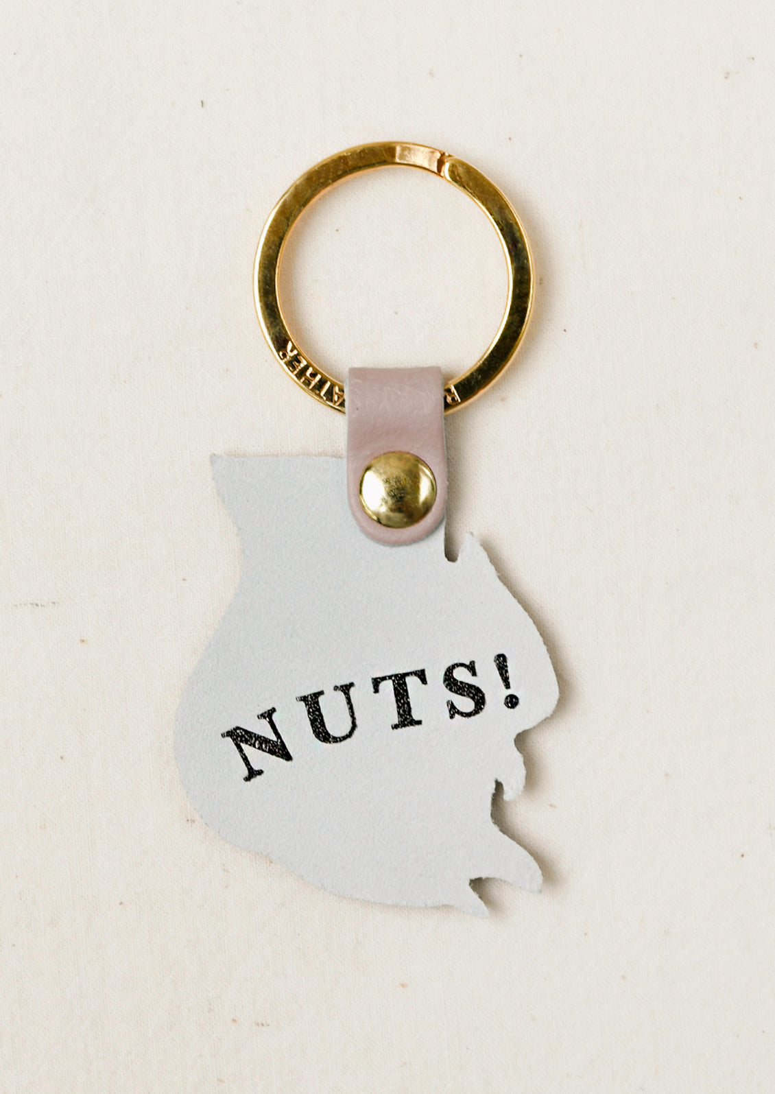 A novelty keychain shaped like a peanut, featuring the word 'NUTS!' printed in black. The keychain has a gold-tone metal ring, a gold-tone rivet, and a pale pink leather strap, all set against a light beige textured background.