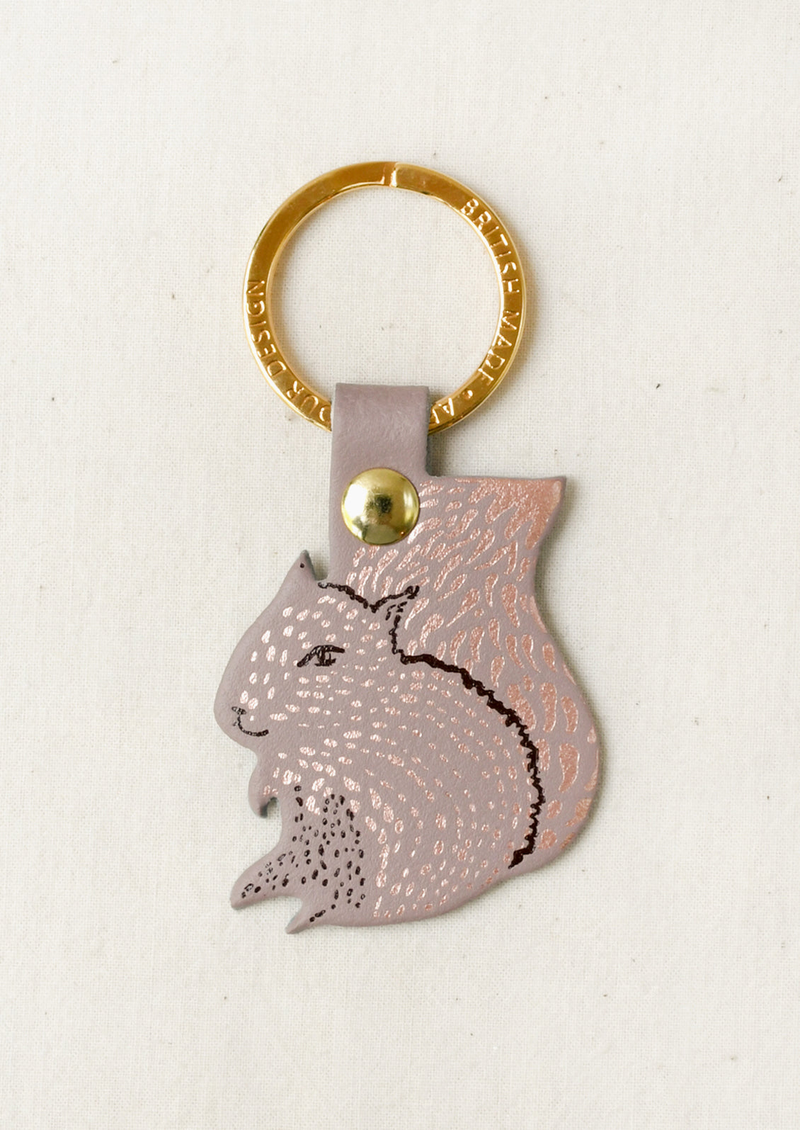 A metallic rose gold keychain featuring a stylized squirrel profile cutout with a gold keyring and a gold rivet, set against a textured off-white background.