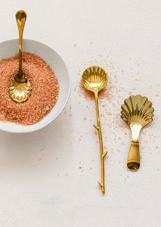 Gold spoons with shell designs on a light surface next to a bowl of pink salt.