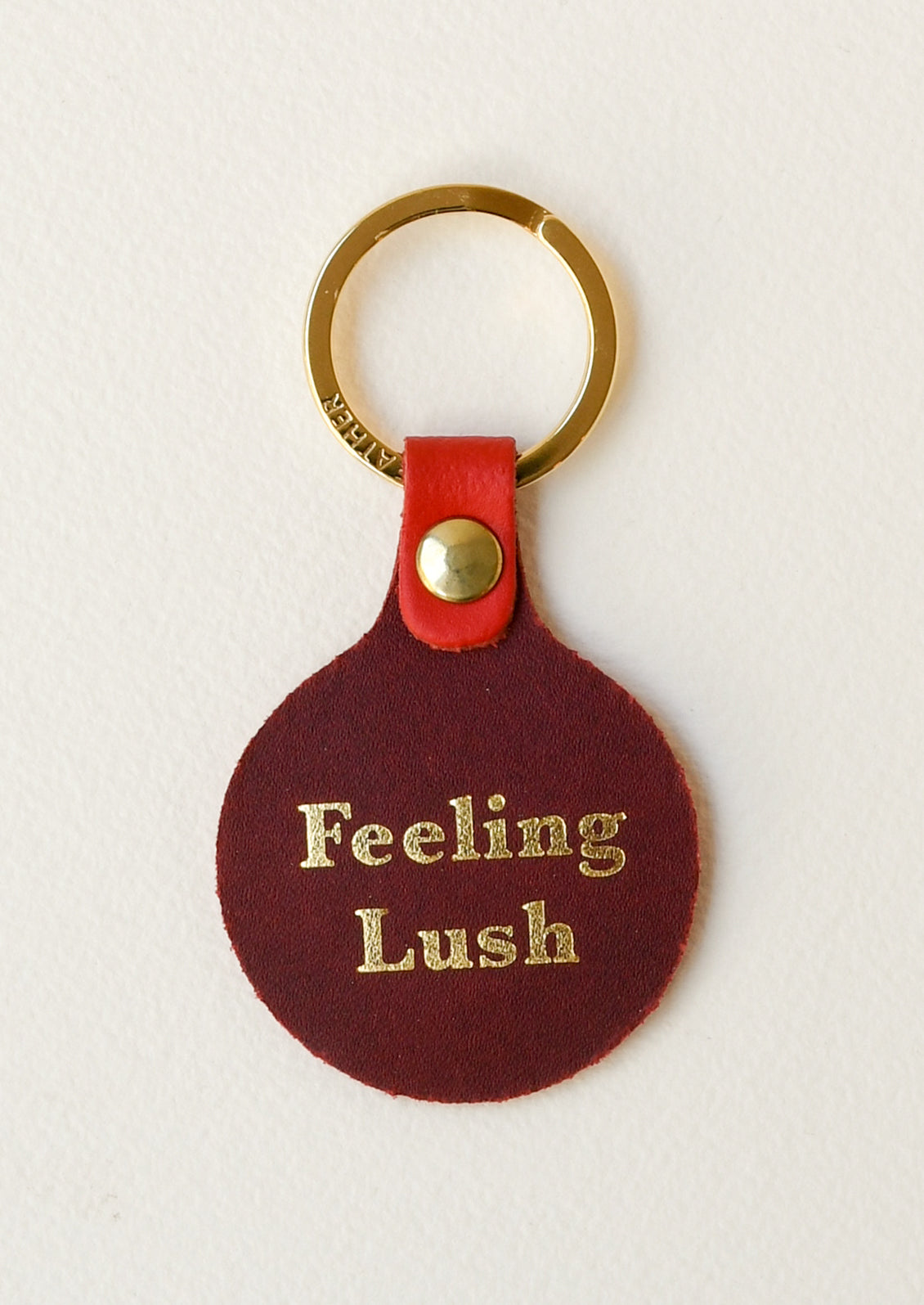 The back of a red leather circle keychain with the words "Feeling Lush" on the back on a gold key ring.