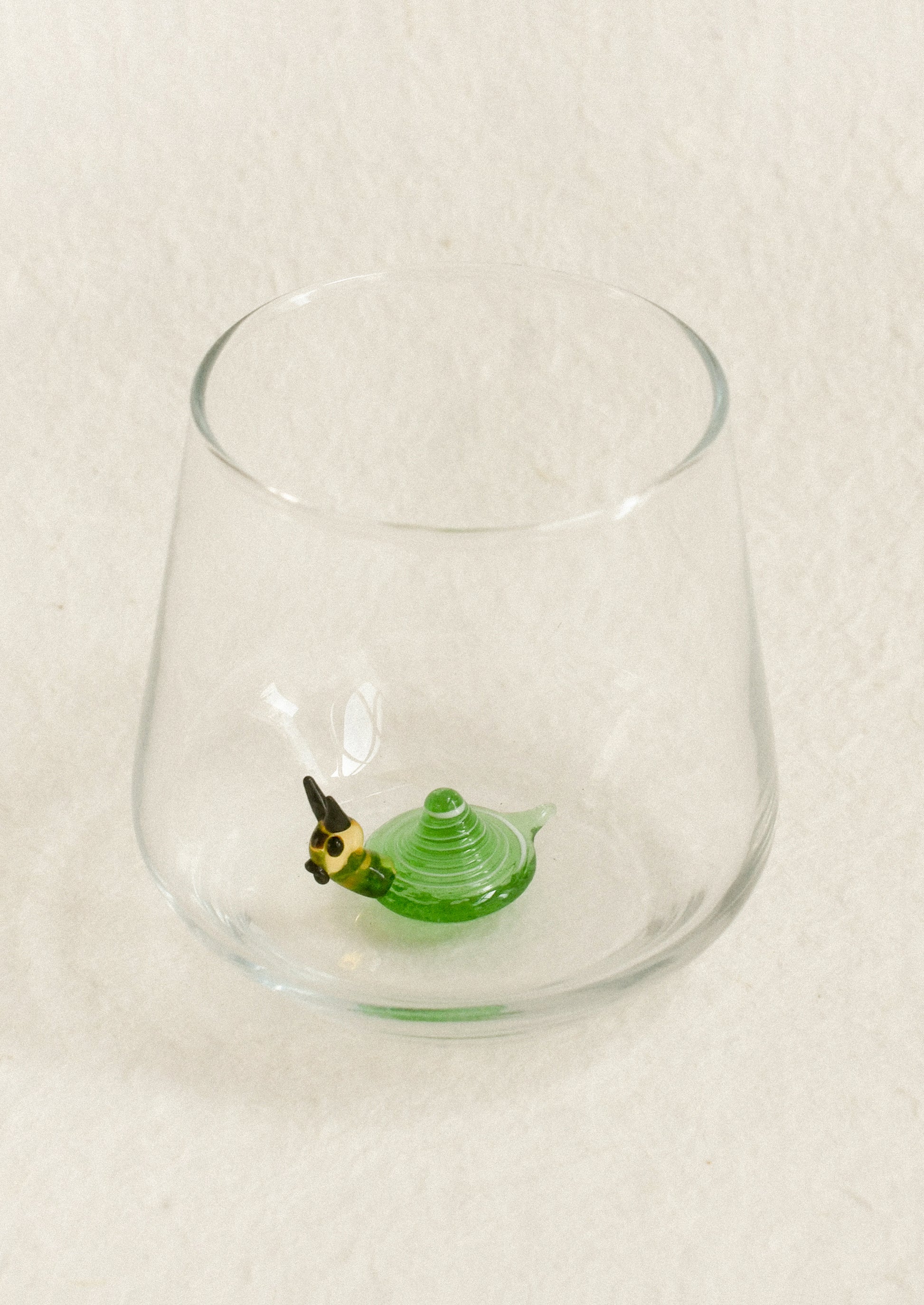 A clear glass tumbler with a small green snail detail near the bottom, photographed from above on a light textured background.