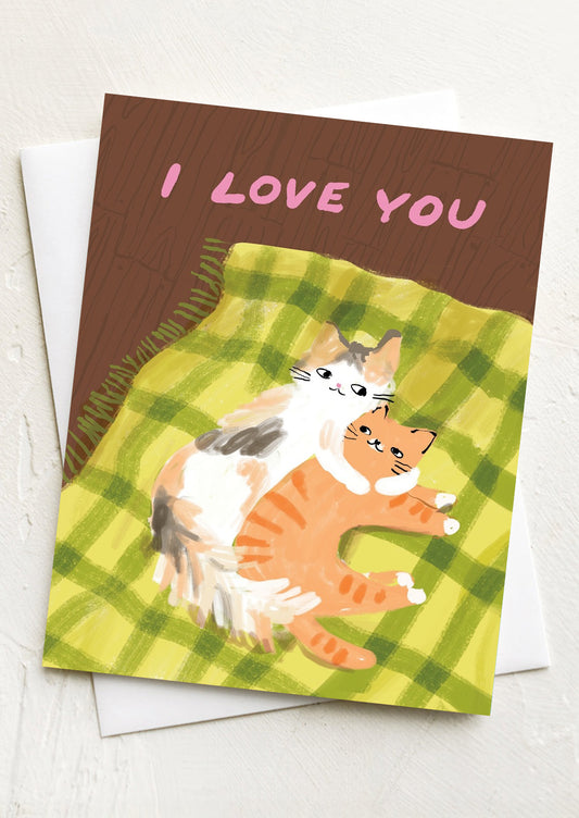 A greeting card with illustration of two cats snuggling in bed, text reads "I love you".