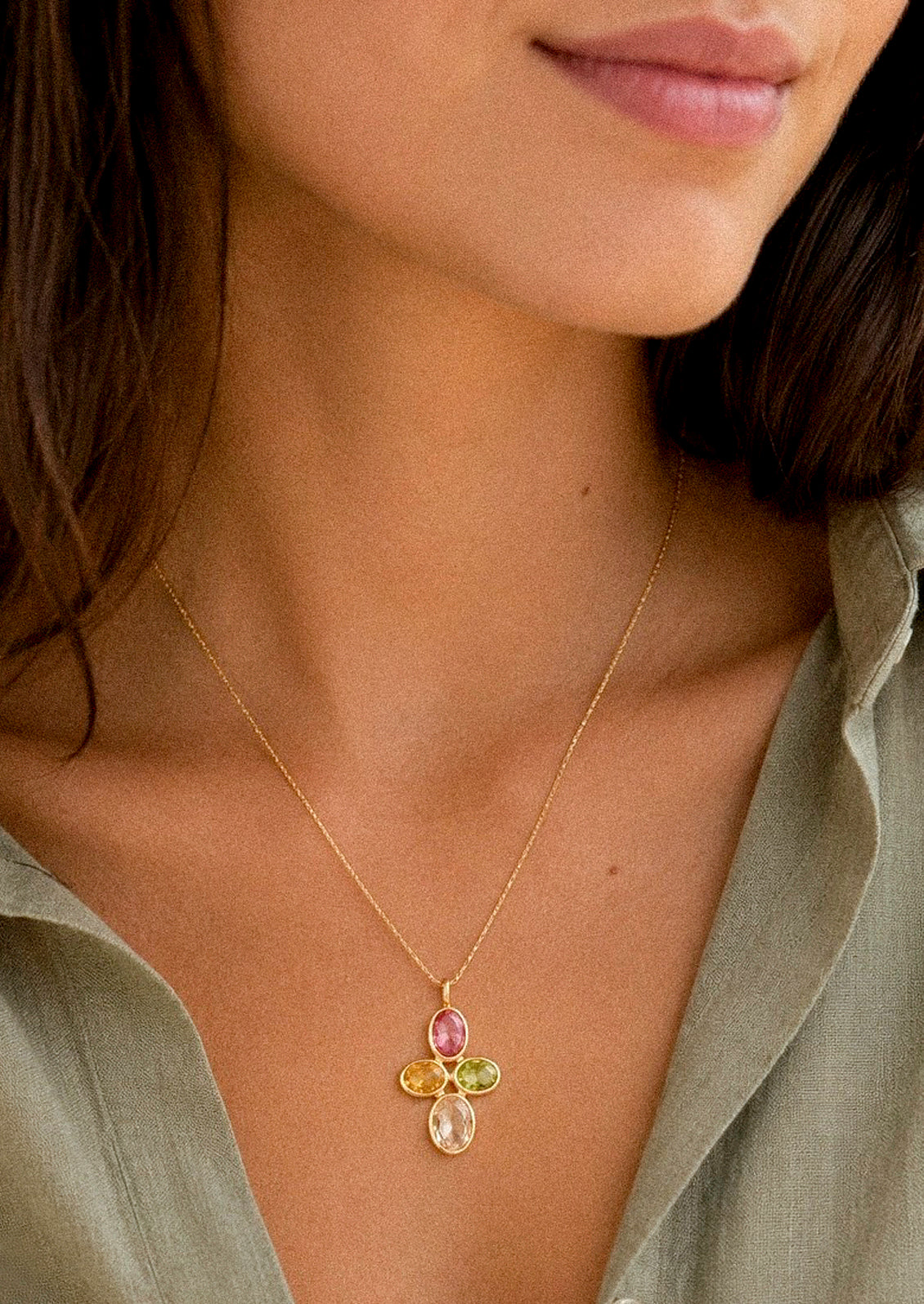 A woman wearing a gold chain necklace with a pendant made with colorful gems in a cross shape.
