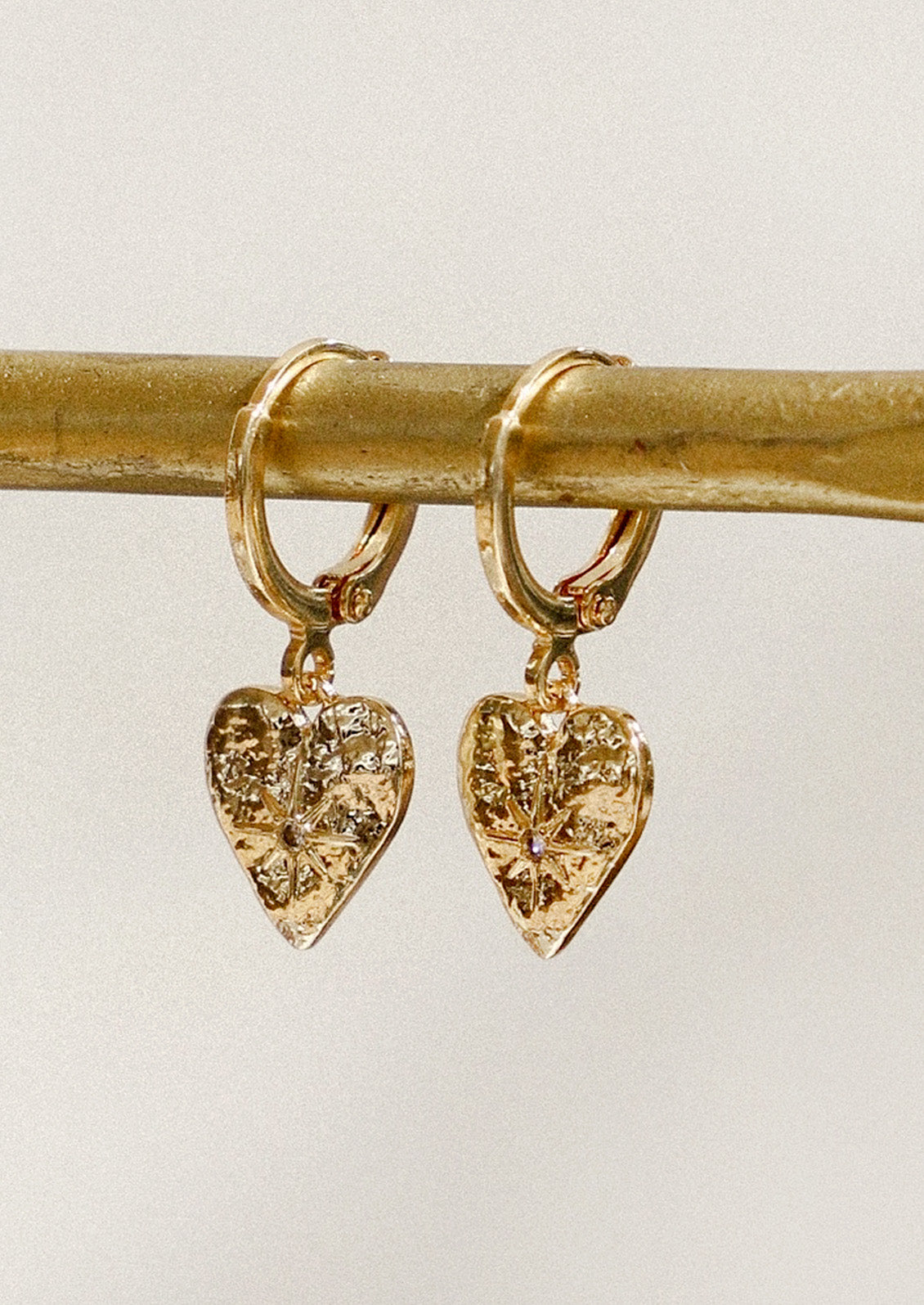 A pair of gold huggie hoop earrings with heart charms with a starburst design hanging from a gold pole in front of a gray background.