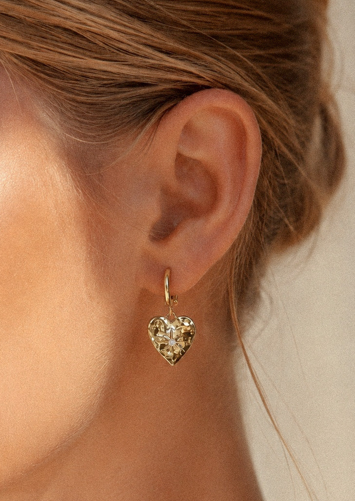 A woman wearing a gold huggie hoop earring with a heart charm with a starburst design and a cubic zirconia gem in the center.