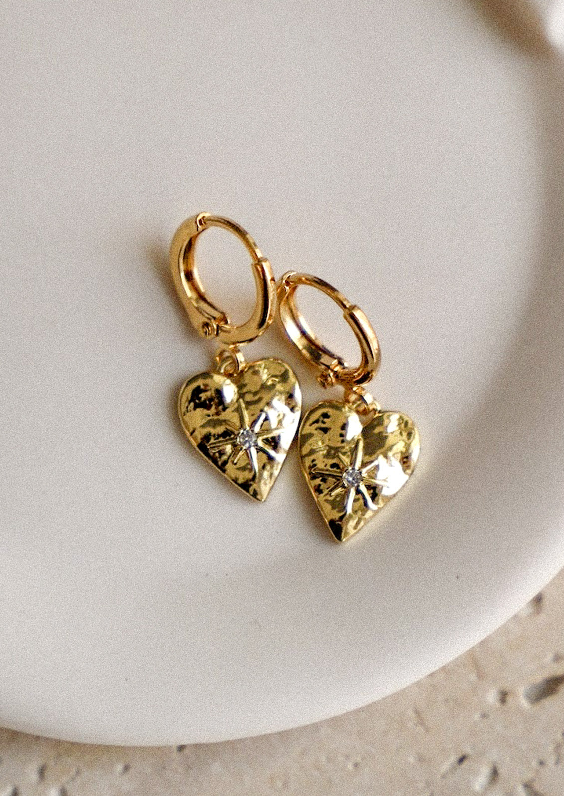 A pair of gold huggie hoop earrings with heart charms and a starburst design with a cubic zirconia gem in the center on a white plate sitting on a stone surface.