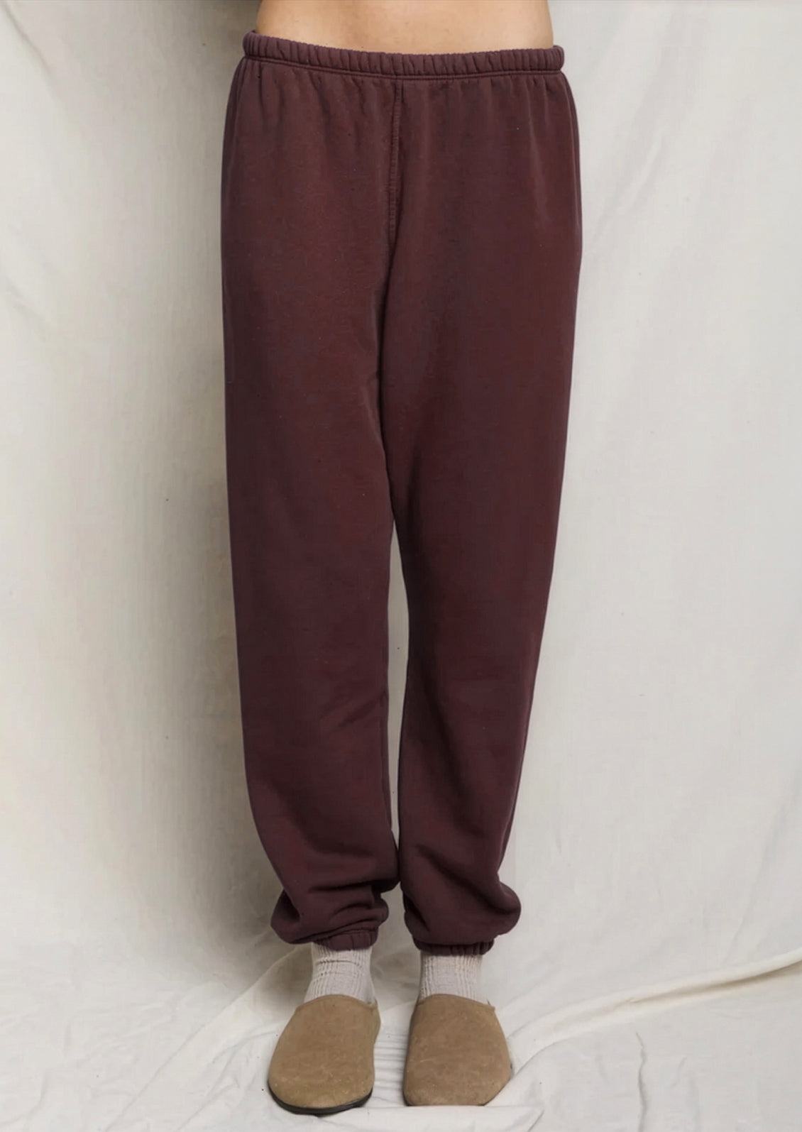 A person wearing loose, dark brown jogger sweatpants with an elastic waistband and cuffed ankles, paired with beige slip-on shoes against a white fabric backdrop.