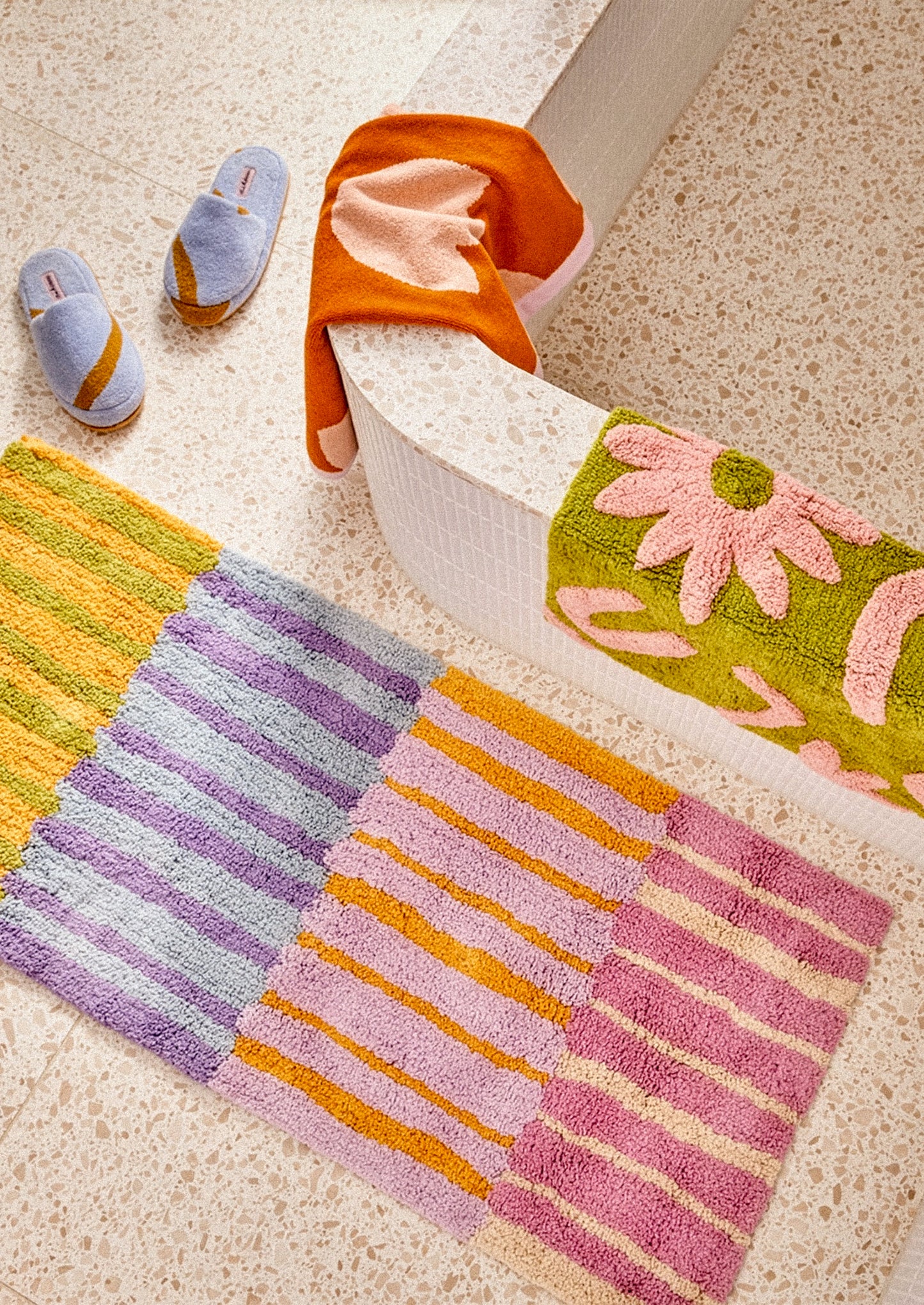 A colorful patchwork bath mat with striped and floral sections in pink, purple, orange, and green, styled on a beige terrazzo floor next to a white tub with an orange towel and slippers.