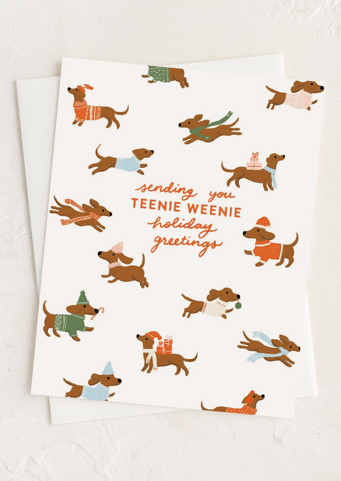 A white holiday greeting card featuring a playful illustration of dachshunds wearing colorful sweaters and scarves, with the text 'sending you TEENIE WEENIE holiday greetings' in orange and brown script. A second white card is partially visible underneath on a light textured background.