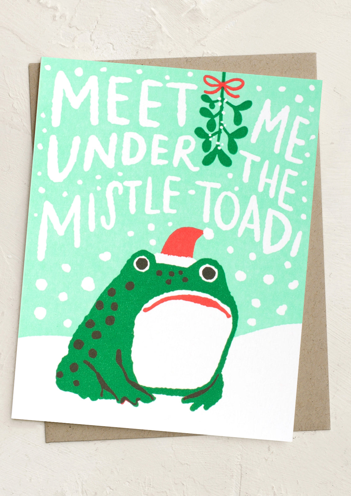 A Christmas greeting card featuring a green toad wearing a red Santa hat, with the text 'MEET ME UNDER THE MISTLE-TOAD!' and a mistletoe sprig on a mint green background, shown with a brown envelope behind it.