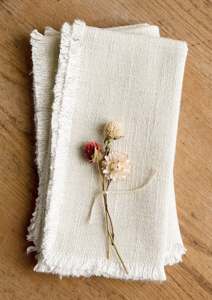 A pair of cream linen napkins with linen texture and fringed edges.