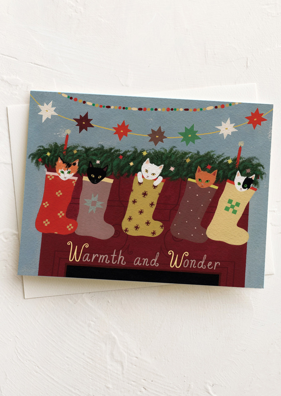 A greeting card with illustration of cats hanging in stockings on a mantel, text reads "Warmth and Wonder".
