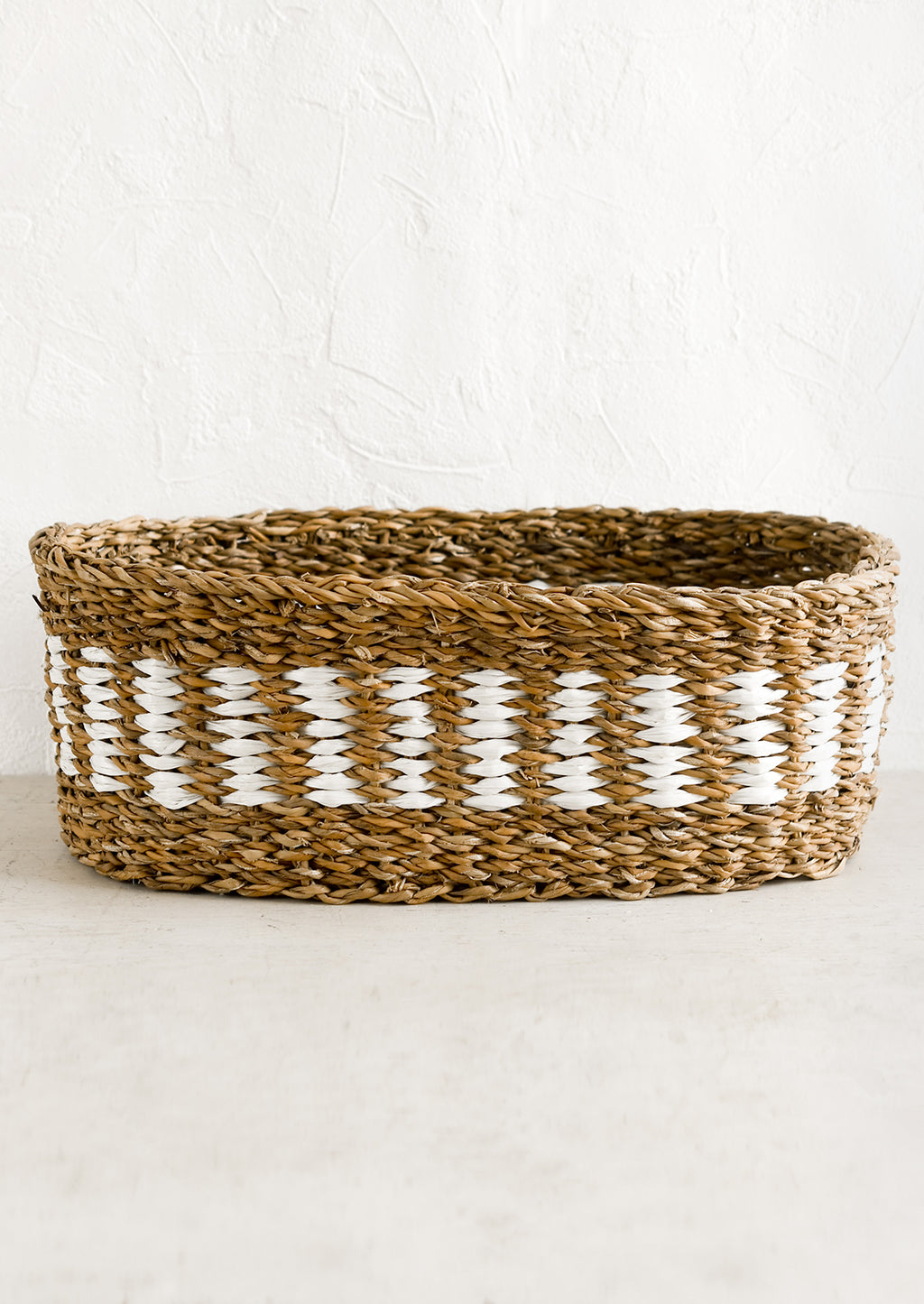 1: An oval shaped storage basket in seagrass with woven white detail.