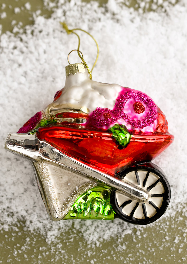 A holiday glass ornament of a red wheelbarrow carrying flowers.