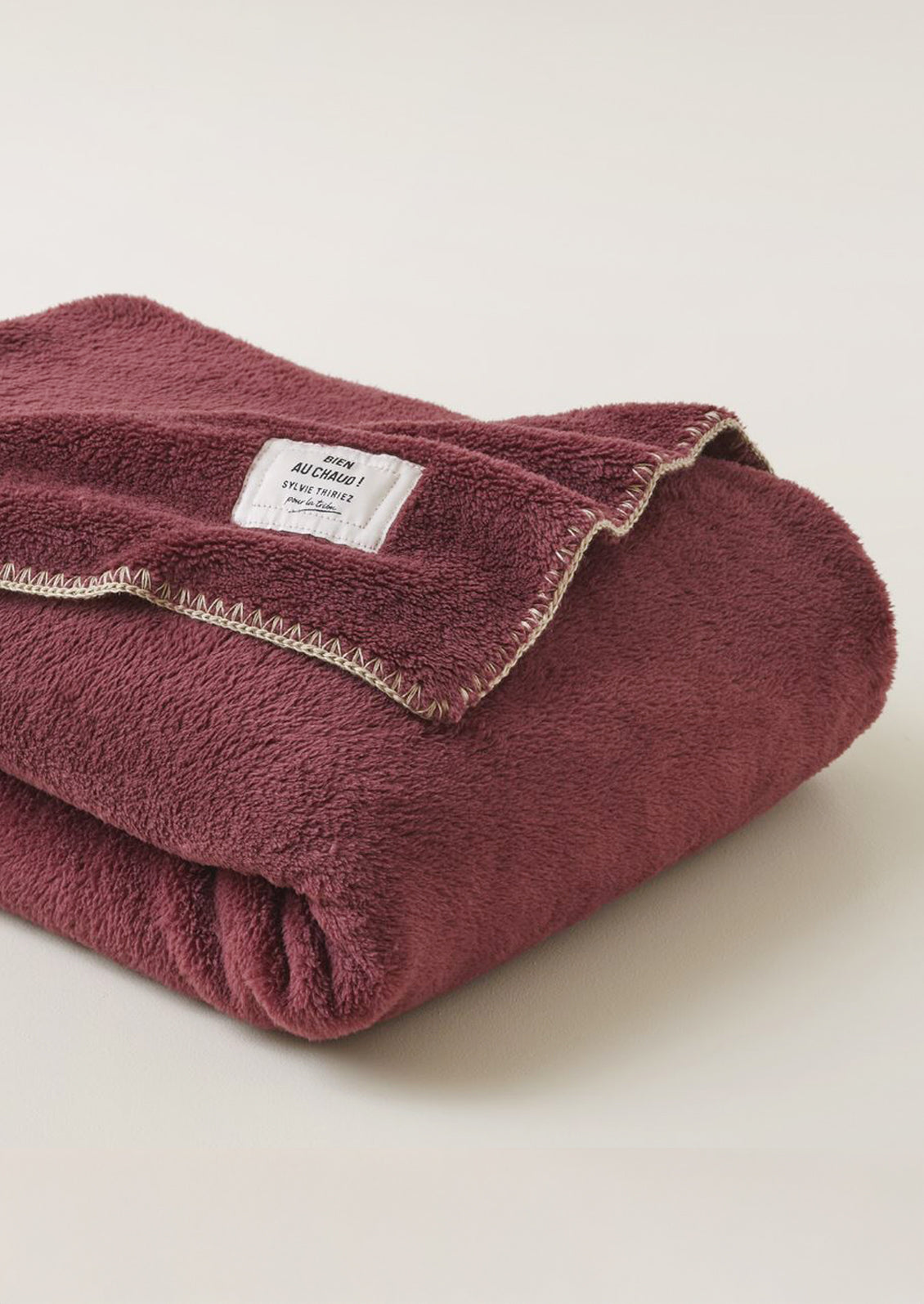 A folded wine-colored plush fleece blanket with a cream-colored stitched border and a white brand label.