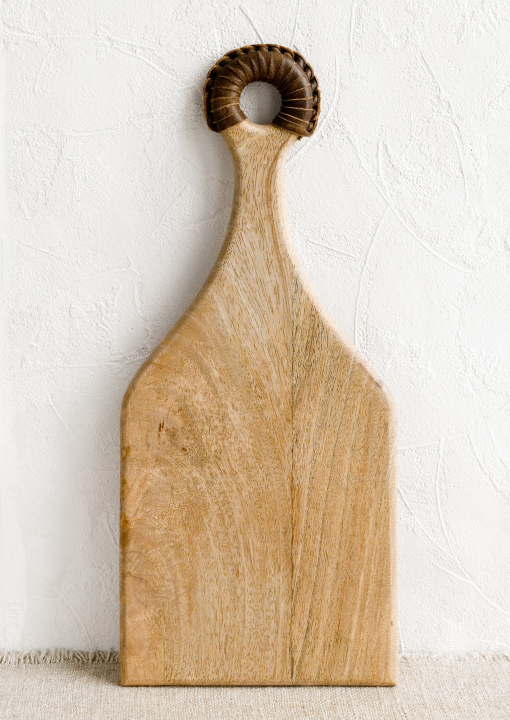 1: A paddle shaped light wood cutting board with brown leather wrapped handle.