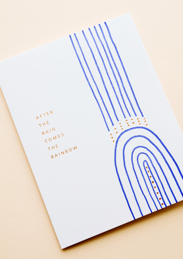 White greeting card with cobalt blue rainbow and gold text reading "After the rain comes the rainbow"