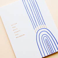 2: White greeting card with cobalt blue rainbow and gold text reading "After the rain comes the rainbow"