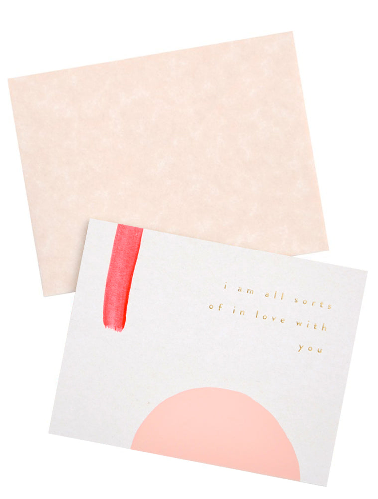 A pale tan envelope and white greeting card with a rectangle of red paint, a semicircle of pink paint, and gold foil text reading "I'm all sorts of in love with you."