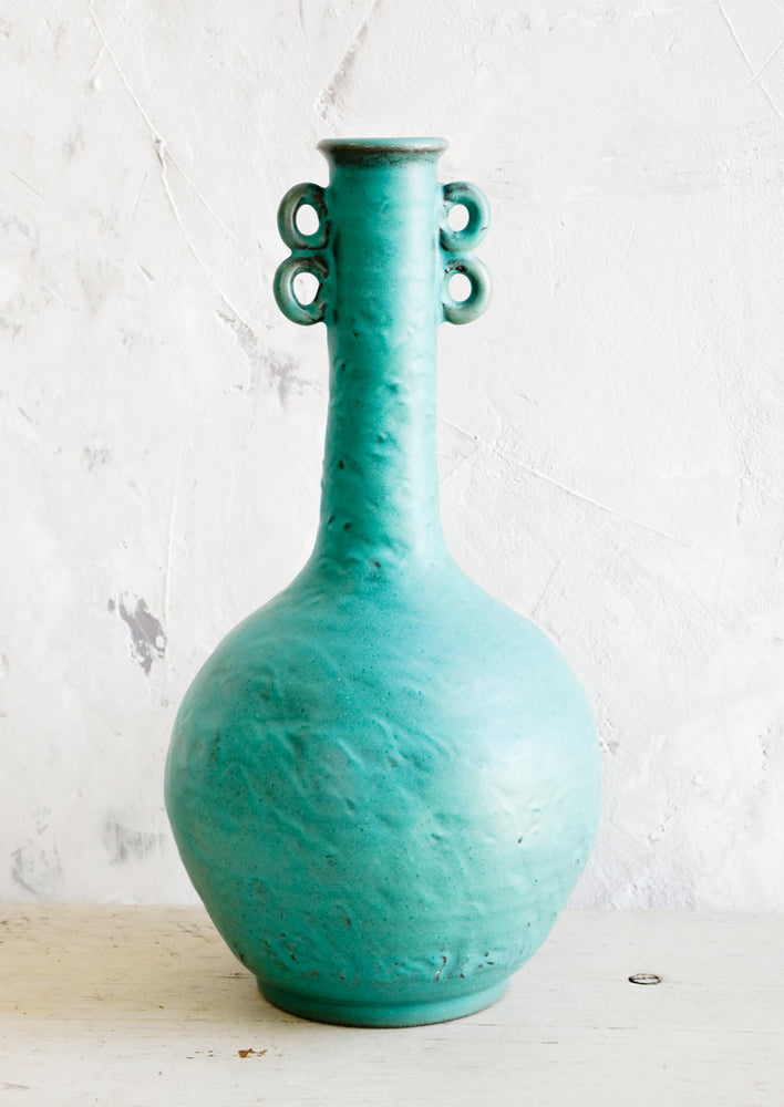 1: Tall, sculptural ceramic vase in turquoise color with bulbous base and skinny neck. Decorative loop detailing at top sides.