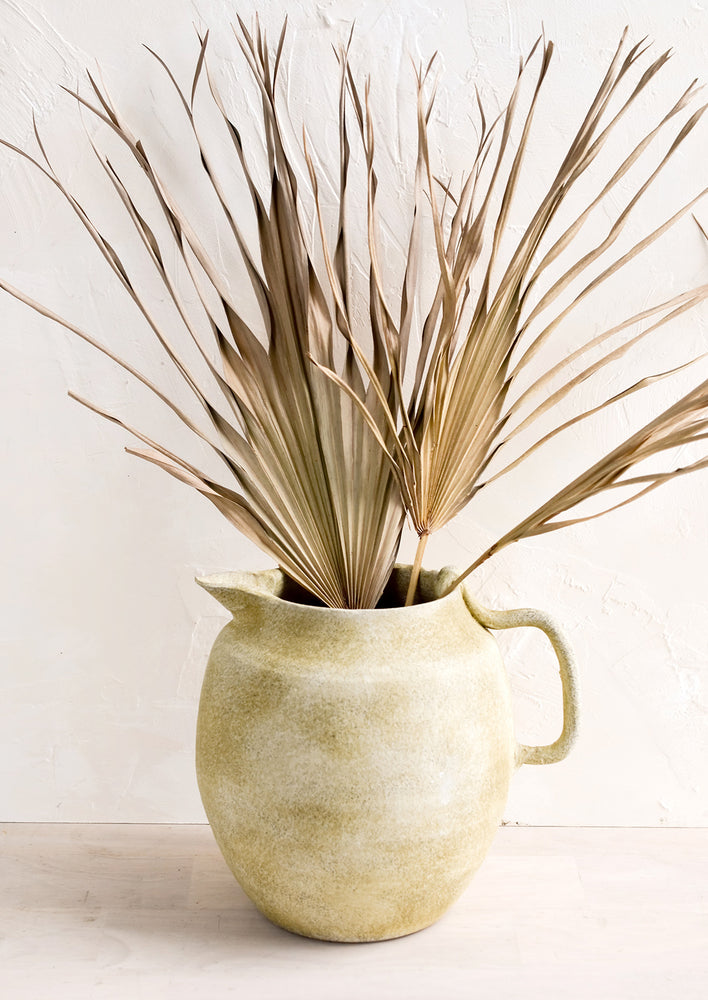 A ceramic pitcher meant to be used as a vase in distressed clay with dried palm leaf.