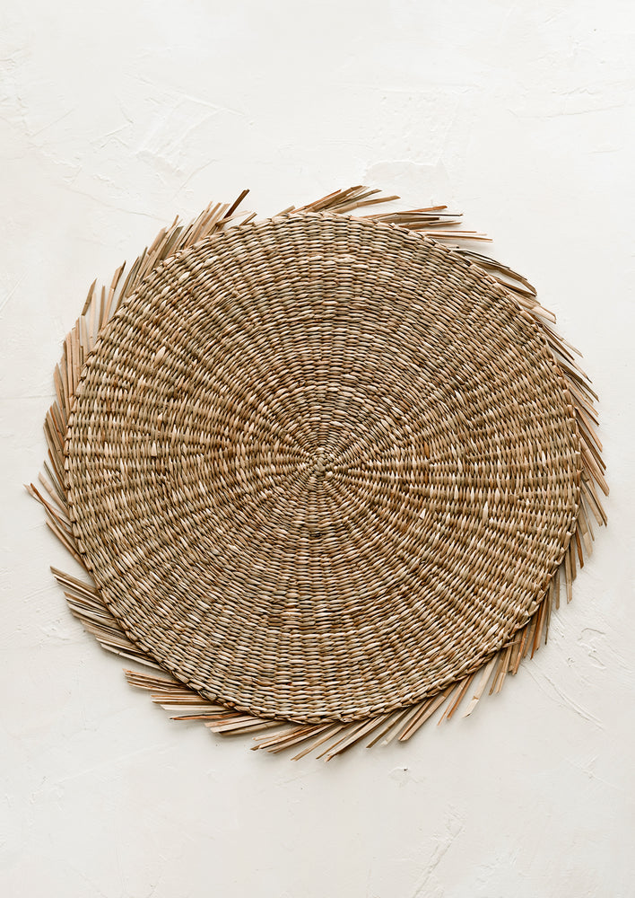 A round straw placemat in natural with curvy fringe trim.