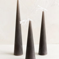 Small / Stone: Three lit cone-shaped taper candles in dark grey.
