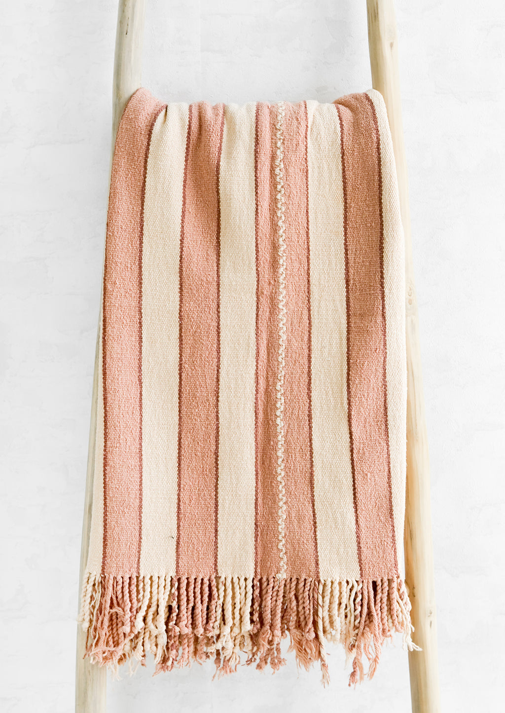 Nude / Blush: Natural woven cotton throw in blush and cream stripes with twisted fringe edge, hung on a display ladder.