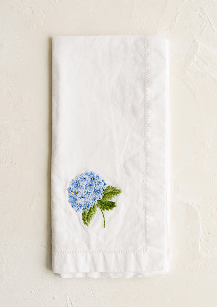 A white cotton napkin with blue hydrangea embroidery.